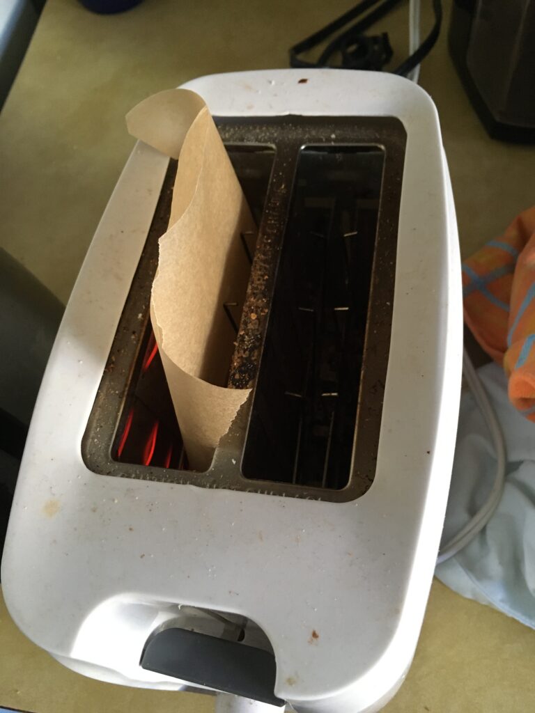 Toaster with two slots, in the left slot is a poptart with a piece of baking foil wrapped around it, the baking foil sticks out.
